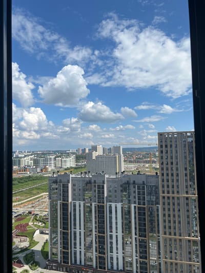 The image captures a panoramic view of a contemporary urban landscape in Yekaterinburg, Russia, under a bright, cloud-dappled sky. In the foreground, two modern high-rise apartment buildings dominate the scene. The building on the left features a striking facade with alternating bands of grey and white, interspersed with vertical panels of reflective glass. Its lower section appears to be a parking lot with parked vehicles and a children's playground with red and yellow play structures, surrounded by green lawns and paved walkways.
The building on the right is taller and exhibits a more uniform design, with a beige exterior and regularly spaced windows. Between these two prominent structures, a network of roads and green spaces is visible, with some areas appearing to be under construction, indicated by the presence of a yellow construction crane in the distance.
Further in the background, a sprawling cityscape unfolds, characterized by a multitude of residential and commercial buildings of varying heights and architectural styles. The cityscape extends to the horizon, where a faint haze suggests the presence of more distant urban development or natural features. The overall impression is one of a rapidly developing and modern city, bathed in the clear light of a summer day.