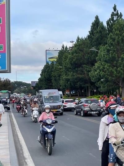 This image captures a busy street scene in Da Lat, Vietnam. A wide asphalt road is filled with a variety of vehicles, predominantly motorbikes, along with cars and vans. The traffic appears to be moderate to heavy, with vehicles moving in multiple lanes.
Several individuals are visible on motorbikes, most wearing helmets and face masks, a common sight in this region. One person in the foreground rides a dark blue motorbike, wearing a pink jacket and a white mask. To their right, another person on a motorbike wears a beige jacket and plaid shorts, also masked. In the mid-ground, other motorcyclists are seen, some with passengers. Cars and vans are interspersed within the traffic, including a silver SUV with a spare tire on the back and a grey minivan.
The environment suggests an urban or semi-urban setting. On the right side of the road, a dense line of tall pine trees forms a natural barrier, creating a green backdrop. Overhead, a cloudy sky is visible, with a hint of blue. In the background, a billboard with a yellow bus graphic is partially visible, and another large billboard with a pink and green advertisement is seen on the left edge of the frame. Streetlights and utility poles are also present, indicating a developed area. The overall impression is one of everyday life and movement in a Vietnamese city.