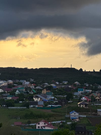 The image displays a panoramic view of a village or residential settlement in Прикамский, Russia, situated beneath a dramatic sky and against a backdrop of a dense, dark green forest.
The sky is a prominent feature, sharply divided by contrasting weather conditions. On the left side, heavy, dark grey storm clouds dominate, creating an intense and brooding atmosphere. In stark contrast, the right side of the sky glows with a warm, golden hue, indicating either a sunset or sunrise, with lighter, more scattered clouds illuminated by the low-lying sun. This dichotomy suggests an ongoing or recently passed weather event.
Below the sky, a continuous ridge of dark green, densely packed trees forms a natural boundary. Perched atop this forested hill, silhouetted against the golden light, is a tall, slender, rectangular structure, possibly a chimney or a communication tower, serving as a distinct landmark on the horizon.
The residential area in the foreground consists of numerous houses, varying in size and design. Their roofs exhibit a diverse palette of colors, including red, blue, green, grey, and brown. Unpaved dirt paths or roads wind through the settlement, connecting the different properties. Various fences, including some made of green mesh and corrugated metal, are visible, marking property lines. No individuals are present, and no specific activities are taking place. The overall impression is a serene yet powerful depiction of a rural landscape under a dynamic sky, highlighting the interplay between human habitation and natural forces. No visible text is present in the image.