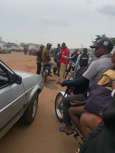 The image captures a bustling street scene in Nyanya, Nigeria, under an overcast sky. The primary subjects are a group of people, many of whom are gathered around motorcycles. A man in a grey shirt and a dark baseball cap with an "ADC" logo is prominently featured in the foreground, leaning forward as if talking or interacting with someone on a motorcycle. Another person, wearing a yellow garment and a dark blue bag, is seated behind him on the same motorcycle.
Several other individuals are visible, some standing and some on motorcycles, suggesting a period of waiting or congestion. A person in a red shirt and khaki pants is also a focal point, standing near a motorcycle. The presence of multiple motorcycles and what appears to be a silver car in the foreground indicates a busy urban or semi-urban environment. The ground is a mix of dirt and asphalt, typical of many Nigerian roads.
The overall atmosphere seems to be one of everyday activity, possibly a market area or a transportation hub where people are waiting for rides or engaging in commerce. The cloudy sky suggests it might be midday, but the low light conditions do not provide definitive cues for the time of day. There are no strong indicators of specific emotions, but the crowd and the proximity of people suggest a social interaction. In the background, glimpses of buildings and more vehicles can be seen, further reinforcing the urban setting. One of the vehicles in the background appears to be a white minibus, commonly used for public transport in Nigeria.