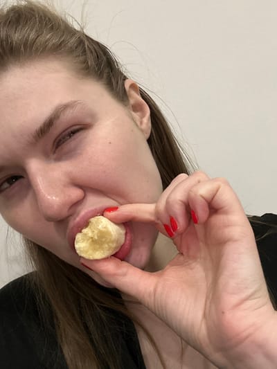 A woman holds a piece of banana to her mouth with her right index finger. Her lips are slightly parted, and her finger presses against the banana. She has light skin, brown hair, and wears red nail polish. The background is plain and slightly out of focus, seemingly a white wall. The composition is unusual, angled with the woman's face tilted.