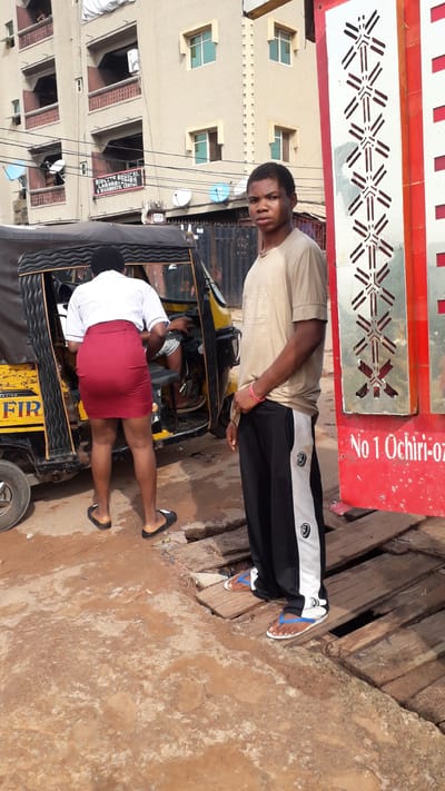 In Nkpor, Nigeria, a young male stands on wooden planks in front of a red sign that reads "No 1 Ochiri-Oz." He wears a beige t-shirt and black pants with white stripes. To his left, a woman in a white shirt and red skirt stands by the open door of a yellow and black auto-rickshaw. The woman is facing away from the camera, and her lower legs and feet, clad in black flip-flops, are visible. The auto-rickshaw has text on its side, including "DELUXE" and what appears to be "FIR." Behind the auto-rickshaw and the people, a multi-story building with balconies and windows is visible, along with various satellite dishes and electrical wires. The ground appears to be a mix of dirt and weathered wooden planks.