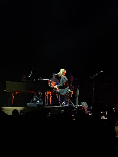 In a dimly lit concert hall, likely in Tbilisi, Georgia, a musician with blonde hair sits at a grand piano, performing under spotlights. He is wearing a dark suit jacket and is looking towards the microphone in front of him. To his left, another person is visible behind the piano, partially obscured. To his right, a person in a suit is playing a saxophone. In the foreground, the silhouettes of an audience are visible, many holding up phones to capture the performance. The stage is decorated with red and blue lighting accents.