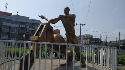 A bronze statue depicts a man and a ram. The man stands upright, bracing himself against a lever or beam that is connected to the ram. Both figures are stylized and somewhat abstract. A white metal fence encloses the statue. The backdrop includes a multi-story building and power lines. The scene is set in an urban area, with greenery visible behind the fence. The sky is clear and blue.