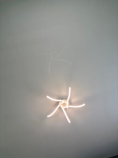 An overhead shot captures a modern, starburst-shaped ceiling light fixture. The fixture emits a warm, white light, illuminating the surrounding grey ceiling. A faint, ghostly reflection of the light fixture is visible on the ceiling above, creating a subtle echo of the main object. The background is an unadorned, textured grey ceiling. The scene appears to be an interior space, likely a room in a residential or commercial building. The specific location is a room in Kableshkovo, Bulgaria.