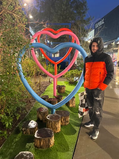 A smiling person in a black and orange jacket is leaning on a large, colorful sculpture with red, pink, and blue curved elements. This structure appears to be part of an outdoor play area with tree stump steps on artificial turf. Behind the person is a store and a checkered shopping bag. The image is taken at night, and the ground appears wet.