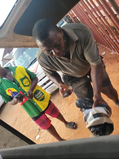 In the city of Awka, Nigeria, a man in a dark olive green t-shirt and dark shorts is seen holding a black object wrapped in grey and white tape, which appears to be a packaged item. He is standing next to another person wearing a green and yellow shirt with red and black striped shorts. The second person is holding a smartphone in their left hand and their right hand is partially visible. The background shows a reddish-brown dirt ground, a white bus with the license plate FGG 133XC visible, and a reddish-orange metal fence. There is also some debris on the ground. The lighting suggests it is daytime.
