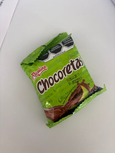 A single, unopened "Chocoretas" candy bar, produced by the Ricolino company, is diagonally positioned on a white surface. The candy wrapper is light green with "Chocoretas" printed in brown and numerous instances of "CHOCOMENTA" and "coretas" in green. A graphic shows a chocolate snack submerged in a chocolate liquid or sauce. A date of 06 JUL 26 is printed on the wrapper. Nutritional information, including warnings about excess calories, sugars, and saturated fats, is presented in a hexagonal design.