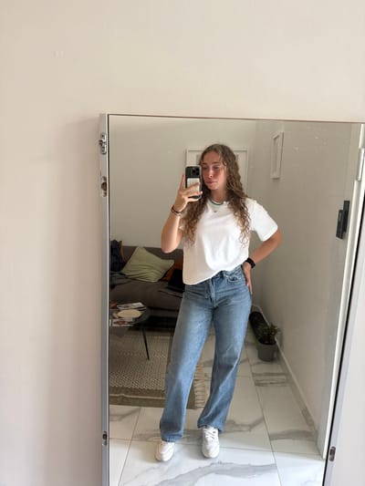 A young female individual is depicted taking a full-length mirror selfie within an indoor residential environment located in Tel Aviv-Yafo, Israel. The individual has long, curly light brown hair and is wearing a white short-sleeved t-shirt, light blue high-waisted jeans, and white sneakers. She holds a smartphone with a white and grey patterned case in her right hand, positioned to capture her reflection, while her left hand rests on her hip. Her attire is accessorized with a thin turquoise beaded necklace, several bracelets on her left wrist, and a black smartwatch also on her left wrist.
The mirrored reflection reveals a room with light-colored walls and a floor composed of white and grey marble-patterned tiles. To the left, a dark grey sofa is visible, adorned with a light green throw pillow and a laptop. A small, black, round coffee table with thin legs is positioned in front of the sofa, holding papers or magazines. To the right, a green skateboard leans against the wall next to a small potted plant housed in a grey pot. A white-framed object is mounted on the wall in the upper right portion of the reflection. The lighting is bright and ambient. The full-length mirror itself features a light metallic frame with visible hardware on its left side.