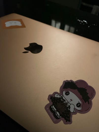 A close-up shot shows the lid of a rose-gold colored laptop with two stickers. A small, light-colored sticker with an orange border is positioned in the upper left corner. Below and to the right of this is the black, iconic Apple logo. A larger, purple sticker featuring a cartoon character, possibly My Melody, is located in the lower right quadrant of the laptop. The character is wearing a brown plaid dress and a black bow. The surrounding environment is dark and out of focus, suggesting an indoor setting. There is no indication of the location being Warsaw, Poland.