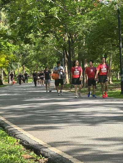 The image captures a shaded pathway in a park, likely in Bangkok, Thailand, given the lush greenery and tropical trees. The path is paved and marked with a yellow line along the edge, bordered by grass and foliage. Several people are walking or jogging along the path. In the foreground, a man in a grey t-shirt and black shorts is looking at his phone while holding an orange bag. Behind him, a group of three people in red t-shirts with race bibs are walking together, two men and a woman. Further down the path, other individuals and groups are visible, some appearing to be participating in a race or organized run. The scene is illuminated by sunlight filtering through the dense canopy of trees, creating dappled shadows on the path.