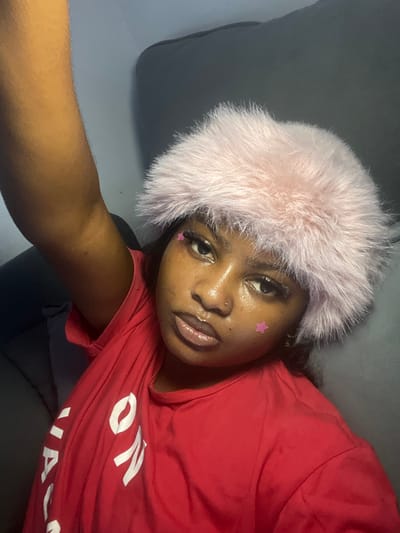 A close-up, high-angle selfie depicts an individual located in Badore, Nigeria. The subject, a person with dark skin, is centered in the frame, looking directly into the camera. They are wearing a pink faux fur hat or headband, a red t-shirt with partially visible white text, and exhibit a small nose piercing on the left nostril. Two small pink star-shaped decals or glitter are affixed to the left cheekbone. The background is dark and out of focus, suggesting an indoor setting with indistinct dark upholstery or furniture. The individual's right arm is partially visible in the upper left quadrant of the image, indicating the act of holding the camera for a selfie.