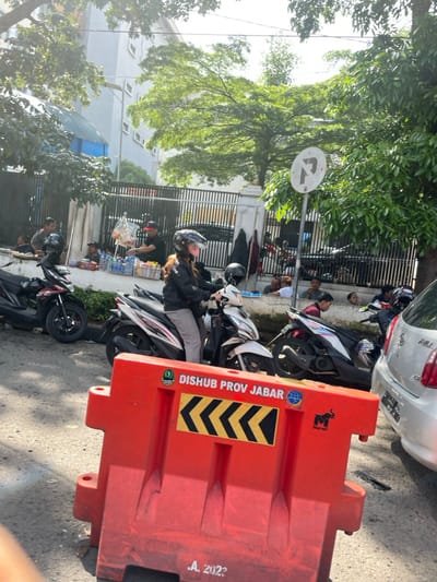A woman wearing a helmet sits on a white and black motorcycle with a black and red motorcycle parked in front of her. Several people are standing and sitting on the sidewalk in the background, including a man with a helmet and a group of people wearing blue and red shirts. There is a large red barrier blocking the street, and a white car is parked behind the barrier. The road is paved and there is a large tree in the background. The scene is taken in the city of Bandung, Indonesia, at the coordinates -6.898 latitude and 107.597 longitude. The barrier is marked with the words "Dishub Prov Jabar" and the year 2022. There is a small elephant emblem on the barrier. The scene is a typical street scene in a busy urban environment in Indonesia.