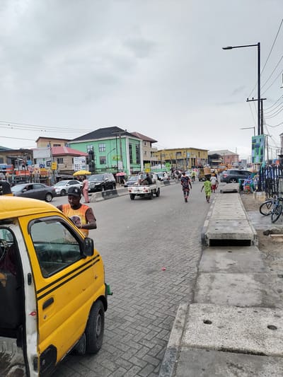 The image depicts a street scene in Lekki, Nigeria. The road is paved with interlocking concrete bricks. A yellow commercial vehicle is parked on the left, with its driver seemingly interacting with someone outside the frame. Several people are walking along the road, some carrying items on their heads. Other vehicles, including cars and a small truck, are driving in the distance. Buildings of various colors and architectural styles line the street. A street light stands tall on the right. The sky is overcast. A concrete drainage ditch runs along the right side of the road with bicycle parked near it.