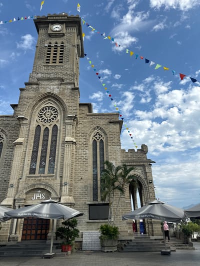 This is a vertical, eye-level shot of the Nha Trang Cathedral in Vietnam. The cathedral, constructed of grey stone with ornate architectural details, is the main subject. A tall bell tower with a clock face is visible on the left side of the image. Colorful triangular flags are strung across the bright blue sky filled with fluffy white clouds, adding a festive touch to the scene. Palm trees and other greenery surround the cathedral. In the foreground, a large, grey umbrella with text printed on it is angled towards the viewer. Part of a paved walkway is also visible. The text on the umbrella reads "Xin giữ thinh lặng, nghiêm trang, trật tự," which translates to "Please keep silent, solemn, and orderly." The overall impression is one of a sunny day at a significant religious landmark.