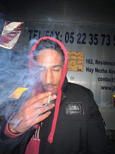 The image features a young adult male photographed in a dark environment, likely at night, against a large, light-colored metallic surface. The male subject, wearing a red hooded sweatshirt beneath a black jacket, has dark hair and a mustache. His right hand holds a lit cigarette or hand-rolled joint, from which a substantial plume of light blue-grey smoke billows, partially obscuring the left side of his face. His eyes are partially closed or squinted. The black jacket has a rectangular patch on the upper left chest, displaying "-18-" and "GDYKJGHUYTR".
The metallic background, consistent with a commercial vehicle (e.g., a truck trailer) or industrial container, is marked with text and graphics. Visible text includes a partial Moroccan phone/fax number "TEL/FAX: 05 22 35 73 5...", an address fragment "162, Résidence Hay Nezha Ain", "contact@", and "www.jl". The "05 22" phone prefix is specifically associated with the Casablanca region in Morocco. "Hay Nezha Ain" is also a residential or commercial area name found within the greater Casablanca region. Additionally, there are industrial pictograms indicating handling instructions (e.g., forklift usage, stacking limits, a "5m" measurement), a "CE" mark, and reflective safety strips: a red and white striped pattern on the upper left and a yellow and black striped pattern on the lower left.
Based on the visible "05 22" phone prefix and the "Hay Nezha Ain" address, the location depicted is indicated as being within the **Casablanca region of Morocco**, not Meknes (which uses different regional phone prefixes such as 05 35 or 05 33). The specific coordinates or detailed street address within Casablanca cannot be further identified from the available information.