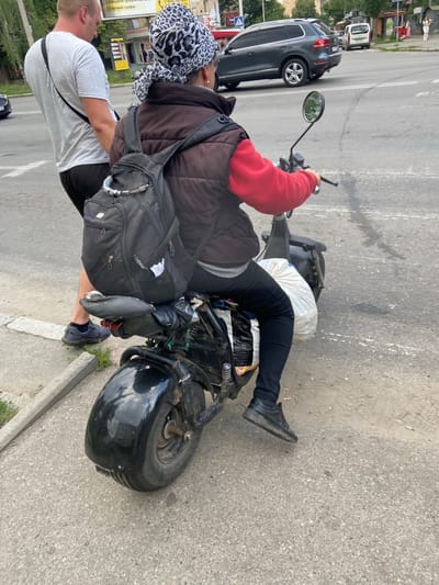 The media file shows two people on a black scooter on an asphalt road, likely in Poltava, Ukraine. The person in the front is wearing a red long-sleeved shirt, black pants, and black shoes. They have a black backpack on their back and a white plastic bag is visible behind them. The person in the back is wearing a brown vest over a darker shirt, black pants, and black shoes. They have a black and white patterned headscarf. A large black backpack is also visible on this person. A dark gray car is parked on the left side of the road, and other vehicles and buildings are visible in the background, suggesting an urban or semi-urban setting. The sky is overcast, indicating possibly cloudy weather. The scene appears to be captured during the daytime. Some text is visible on a yellow sign in the background, but it is mostly illegible.