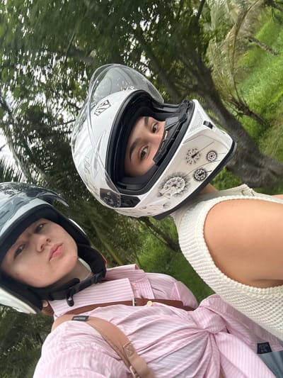 Two individuals wearing motorcycle helmets are positioned closely together in an outdoor environment with lush green foliage and trees in the background, suggesting a tropical location such as Rawai, Thailand. The individual in the foreground wears a black and gray helmet and a pink and white striped shirt, with a brown leather strap across their chest. Their gaze is directed towards the camera with a slightly pursed expression. The individual behind them wears a white helmet adorned with a dandelion-like graphic and is looking upwards, with their right eye visible through the helmet's visor. Their upper body is clad in a cream-colored knitted top. The scene appears to be captured from a moving vehicle, possibly a motorcycle, given the tilt of the image and the framing of the individuals. The overall atmosphere is casual and dynamic, suggesting a shared activity or journey in a natural setting.
