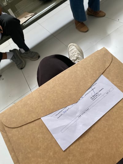 A close-up, overhead shot shows a person's legs and feet, one in black pants and gray sneakers, the other in dark brown pants and white sneakers, sitting with a large brown envelope on their lap. The envelope contains a document with visible text. The document shows a signature and the name "DR. KRISTIANN J. CABONITA," along with "Medical Officer III" and "License No. 0135115." It also mentions "Davao City, Philippines" and "Health Services Division." The background reveals a tiled floor and a metal bench, suggesting an indoor waiting area, likely in Davao City, Philippines.