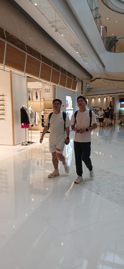 Two young Asian males walk down a brightly lit, modern interior hallway. The male on the left wears glasses, a white t-shirt, white shorts with red detailing, and light-colored, patterned high-top sneakers. He has a black backpack. The male on the right wears a light pink t-shirt, dark pants, and white sneakers. He also has a black backpack and is holding a smartphone in his right hand. They are walking towards the viewer, and the background shows a clothing store with mannequins and displays. Further in the background, other people are visible near escalators and store entrances. The floor is highly reflective and appears to be polished white tile. The location is identified as Jiulongpo District, China.