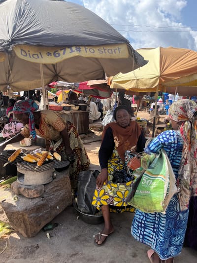The image depicts a vibrant outdoor market scene in Gwong, Nigeria. In the foreground, two women are engaged in selling and preparing food. One woman, wearing a colorful yellow and black patterned dress, is seated and appears to be resting. The other woman, in the lower half of the image, is tending to corn being grilled over an open fire. She wears a patterned headscarf and a dress with a geometric design. The grilling setup includes a metal rack over what appears to be a makeshift stove or brazier, with several ears of corn cooking.
Behind the women, the market unfolds with various stalls and vendors. Numerous umbrellas and awnings provide shade. One large umbrella, predominantly gray and khaki-colored, has a yellow band with purple text. Partially visible text on this band reads "Dial *671#. To S". Another green bag, with visible text "50kg NET WEIGHT" and a phone number, is placed near the seated woman.
The scene is bustling with activity, suggested by the presence of many people and a variety of goods on display, though not all are clearly discernible. The lighting indicates it is daytime, likely with sunny weather given the strong shadows and bright sky visible in the upper left portion of the image. The overall impression is one of lively commerce and everyday life in a Nigerian market.