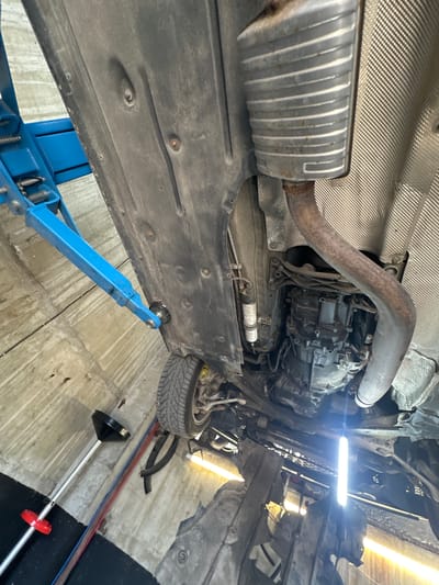 An automotive repair shop in Kazanlak, Bulgaria, contains a car elevated on a blue hydraulic lift. The underside of the vehicle is visible, revealing the exhaust system and transmission. A technician's tool, likely a funnel on a wand, is on the floor to the left. The scene is lit by overhead shop lights, creating a somewhat dim environment.