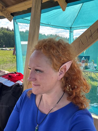 An individual with reddish-brown curly hair and prosthetic elf ears is observed in a profile view under a wooden and blue tarp canopy in Большая Кивара, Russia. The person, appearing female, is looking towards the left of the frame with a subtle smile. She wears a royal blue long-sleeved top and a black cord necklace with a green pendant. Dark eyeliner and possibly false eyelashes are visible. The canopy structure features natural wooden beams; one beam displays two hand-drawn hearts, while another has black markings resembling "Л 14". In the immediate foreground to the left, a red and black item, possibly a jacket or bag, is present. The background reveals a bright outdoor setting with a clear sky and scattered white clouds over a large green grassy field bordered by dense green trees. Further in the background, underneath the canopy to the right, a small table holds two bottles (one containing a red liquid, another a yellowish liquid) and some plastic bags. In the far left background, a red, white, and blue object, consistent with a Russian flag, is partially visible on the ground.