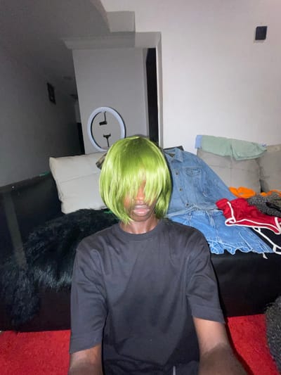 An indoor photograph, taken in Ado, Nigeria, captures a person with dark skin wearing a bright neon green wig that covers most of their face, with only their lips visible. The individual is dressed in a black t-shirt and appears to be lying down or lounging on a sofa, with a white pillow supporting their head. Their right arm is visible, bent at the elbow.

The background reveals a domestic setting, likely a living room. A grey sofa is partially obscured by a pile of various clothes, including a light blue denim jacket, a red garment, an orange fabric item, and a light green and white striped shirt. A fluffy black blanket or rug is draped over a dark furniture piece, possibly a table or armrest.

On the light-colored wall to the left, a circular white decorative item featuring abstract black symbols, resembling a stylized face or characters like '¬ L', is displayed. A small dark square object is also mounted higher on the wall. The room is dimly lit, suggesting the photograph was taken during the evening or at night. The scene has a casual and somewhat playful tone, possibly hinting at a costume or a relaxed moment.