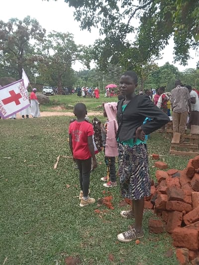 The image shows a group of people standing in a grassy area in Malava, Kenya (latitude: 0.455, longitude: 34.799). A woman with a black jacket and a patterned skirt stands near a pile of red bricks in the foreground. She is looking to the left, and her right hand is on her hip. Behind her, a young girl in a pink jacket and a boy in a red shirt are standing. In the background, there are more people gathered near a white truck. A red cross flag with the words "EKISOBOLA SABASITO" and "YANICA" can be seen in the foreground, indicating a possible medical or emergency response. The people in the background appear to be engaged in some activity, likely a community event or gathering. Trees and foliage surround the grassy area, and the sky is overcast with light clouds.