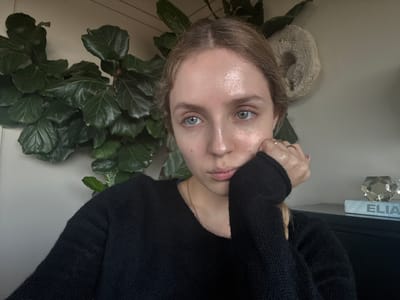 The image is an upside-down close-up portrait of a young woman within an indoor setting. She has fair skin, light brown hair, and striking blue-green eyes that gaze directly at the viewer with a neutral expression. She wears a black knit sweater and a delicate golden chain necklace. Her right hand, adorned with a ring on the ring finger, is held up to her chin. Her forehead shows a slight sheen.
Surrounding her, elements of the room are visible. To her right (if the image were upright), a lush houseplant with large, dark green leaves, resembling a Fiddle Leaf Fig, fills a significant portion of the frame. To her left, near her head, rests a textured, light-colored, possibly ceramic or woven, circular decorative object. Further back on the left side, an object that appears to be a book or block is visible, displaying the upside-down letters "ELIA". Adjacent to this, a clear, multi-faceted crystalline or glass decorative item catches the light. The background is a plain, light-colored wall. The image gives no specific visual cues to the provided location context of Mexico City, Mexico.