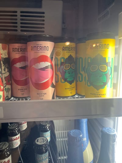 This media file shows the inside of a refrigerator stocked with alcoholic beverages, likely in a store or a home. The primary subjects are various bottles and cans of beer.
On the left side, three cans are stacked vertically. The top two cans are yellow and feature a green cartoonish ape wearing sunglasses, with text indicating "AMERINO BREWING COMPANY" and "GUESSWHAT SESSION IPA." The bottom can is pink and displays a graphic of red lips with a pink tongue sticking out, also labeled "AMERINO BREWING COMPANY" and "BIRRA EDWIGE BLANCHE," with a 4.0% ABV. Below these cans, another can with similar graphics is partially visible.
On the right side, several blue-wrapped bottles are visible, along with some darker bottles with labels that are partially obscured or difficult to read. Some of these labels appear to have Italian text, such as "Daladi ARTIGIANALE" and "QUERA."
The setting appears to be a dimly lit refrigerator, as indicated by the overall dark tone and the reflections on the glass doors. The cool, artificial lighting suggests it could be any time of day, and the location cues from the Italian text on some labels point to Italy. No people are visible, and the objects themselves convey no specific emotions. The overall impression is a commercial display of various craft beers.
