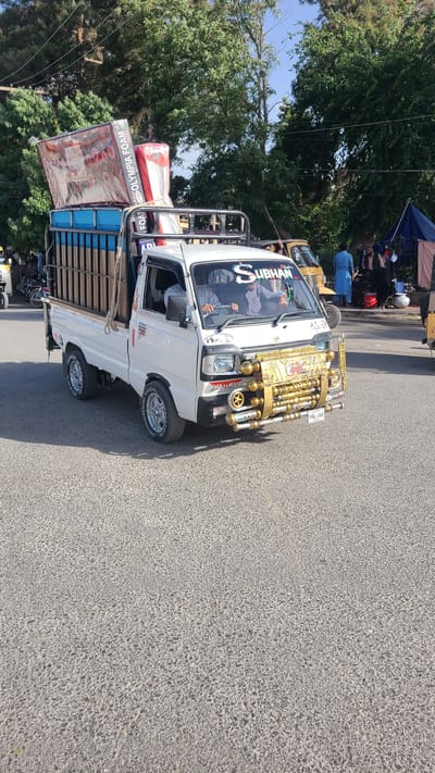 A white mini-truck, likely a pickup or cargo van, is depicted stationary or moving slowly on an asphalt road under bright daylight. The vehicle is heavily customized, particularly at the front, featuring an elaborate gold-colored decorative grill, chrome-plated pipes, additional lights, and what appear to be air horns. A license plate displaying "PAL-349" is visible below the golden decoration. The word "SUBHAN" is prominently displayed in stylized lettering on the top of the windshield.

The driver is visible through the front windshield and side window, appearing to hold the steering wheel. The vehicle's side has some text, possibly "STYLIO" or similar, below the window. The tires show "CROSSWIND RADIAL" branding. The bed of the truck is loaded with several mattresses or foam blocks, one clearly bearing the brand name "OLYMPIA FOAM." The cargo is secured by ropes and sits within blue-painted wooden side rails.

In the background, lush green trees line the roadside, and a clear blue sky is visible. Further down the road, there are other vehicles, including what looks like a yellow auto-rickshaw and motorcycles. Several people are scattered in the background, some near a blue canopy or tent. The overall scene suggests a bustling urban or semi-urban environment in good weather. The image is rotated 90 degrees counter-clockwise from its natural orientation.