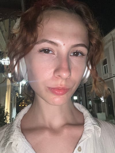 The media file is a close-up, sideways selfie of a young woman with reddish-brown curly hair and light skin. She is wearing white headphones and a textured white shirt with a button closure. Her eyes are brown, and she has on mascara and lip gloss. A light blueish streak of light appears to be reflecting on her face, possibly from a light source. The background is dark and appears to be an urban setting at night, with blurred building lights and structures visible. Hints of greenery are also present. The overall impression is of a casual, personal photograph taken outdoors in a city at night. The provided location context is Novi Sad, Serbia, which could imply the setting is within that city. No visible text is present in the image.