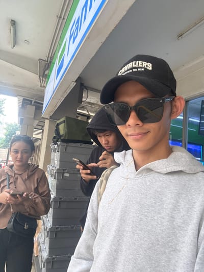 The image captures three individuals standing outdoors, with a FamilyMart convenience store sign visible in the background, suggesting the location is in North Jakarta, Indonesia. The foreground is dominated by a young man wearing a grey hoodie and a black baseball cap with the word "CAVALIERS" and "MAINTAIN THE HEAT" embroidered on it. He is also wearing large black sunglasses and looking directly at the camera, seemingly taking a selfie.
To his left and slightly behind him, another young man in a black hoodie is looking down at his mobile phone. Further to the left, a young woman in a brown hooded jacket and black pants is also looking at her mobile phone. She has her hair pulled back and is wearing a small black crossbody bag.
Behind the people, stacks of grey plastic storage containers are visible, along with a green duffel bag placed on top of some of the containers. The environment appears to be a sidewalk or an area adjacent to a building. The lighting suggests it is daytime.