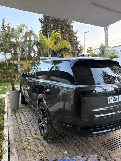 A dark grey Range Rover SUV is parked on a herringbone-patterned brick paver driveway within a residential area of Meknes, Morocco. The vehicle is being charged, with a bright yellow cable connected to a visible port on its front left fender. The Moroccan license plate "64457 I" is affixed to the rear of the vehicle. To the left of the vehicle, a tall, grey cylindrical planter contains a single, dormant plant stalk. The surrounding environment includes numerous palm trees and tall conifer trees, a light-colored wall partially covered with climbing vegetation, and a white overhead structure that extends into the frame from the top. A metal drainage grate is visible on the right side of the driveway. The scene is illuminated by natural daylight. No individuals are visible.