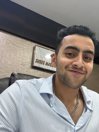 A young adult male, fair-skinned with short dark hair and a light beard, is depicted from the chest up in an indoor commercial setting. He wears a light blue and white vertical striped collared shirt and a silver-colored cuban link chain necklace, maintaining direct eye contact with a subtle smile. The background features a light beige textured wall with gold speckles. Mounted on this wall is a rectangular sign with a gold-colored frame, displaying the partial text "SHREE MAR" in raised, metallic-colored lettering against a light, possibly marble-patterned, surface. Above the textured wall, a dark brown or black horizontal panel runs along the top. A black leather office chair backrest is partially visible on the left. The scene is located within a commercial establishment in Kolkata, India; the partial text "SHREE MAR" on the sign is indicative of common business naming conventions in the city.