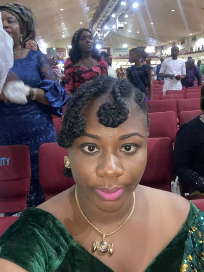 A medium-shot image captures a woman in a green sequined dress, turned towards the camera with a slight upward angle, her right side more prominently displayed. She is wearing a gold chain necklace with a decorative pendant. Her hair is styled with cornrow braids that are gathered into two round clusters at the front of her head. She is wearing pink lipstick. The background is filled with rows of red seats and a crowd of people, suggesting an event or gathering is taking place. People are dressed in a variety of outfits, including a man in a white shirt in the background. The photo may have been taken during an event or ceremony in Ota, Nigeria.