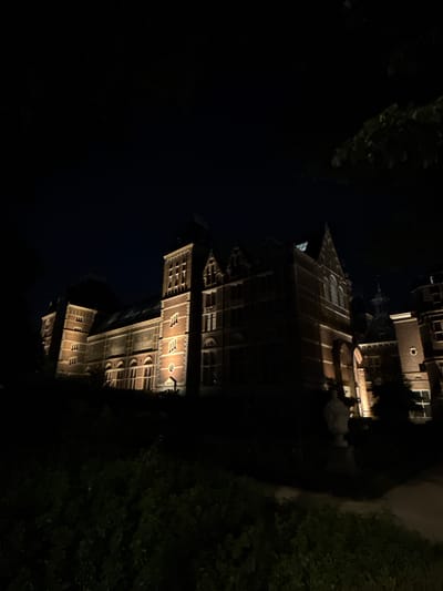 The image shows the Rijksmuseum in Amsterdam at night. The museum, a large brick building with multiple towers and arched windows, is illuminated by warm outdoor lighting, highlighting its architectural details. Dark foliage and trees partially obscure the lower parts of the building and frame the scene from the foreground. The sky above is dark, indicating nighttime. There are no people or vehicles visible in the image.