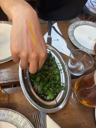 A person's hand with red and yellow streaks of paint on the back of it is reaching into a metal bowl filled with green peppers, likely Padrón peppers, sprinkled with salt. The bowl is placed on a wooden table, alongside white plates with black and white striped rims, cutlery, and a glass with a beverage. The scene appears to be a casual dining setting, possibly a restaurant in Milan, Italy, suggested by the presence of tableware and drinks.