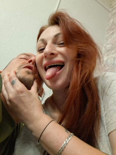 The image shows a close-up, low-angle shot of two individuals. A woman with long, reddish-brown hair is in the foreground, her face angled towards the camera. She is sticking her tongue out and her mouth is open wide, revealing her teeth and gums. She appears to be wearing a light-colored, textured top. A man, partially obscured, is to her left, his face turned away from the camera. His eyes are closed, and his head is tilted back slightly. His hand is touching the woman's face, near her cheek. He has a short beard and a tattoo is visible on his hand. A black string bracelet is worn on the woman's left wrist, and a silver chain bracelet is visible on her right wrist. The background is a plain, light-colored wall. The scene suggests an intimate or playful moment between the two individuals. There is no discernible information in the image to indicate a specific location within Забалоцкі сельскі Савет, Belarus.