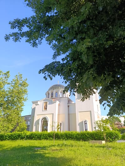 The image shows a white church with a green dome, situated in Razgrad, Bulgaria. The church is partially obscured by lush green trees and foliage, suggesting a natural setting. The sky is clear and blue, indicating a sunny day. The foreground features a well-maintained lawn with some scattered twigs and a small sapling. In the background, an apartment building and other structures can be seen, indicating an urban environment. The lighting suggests it is daytime, possibly late afternoon due to the long shadows cast by the trees.