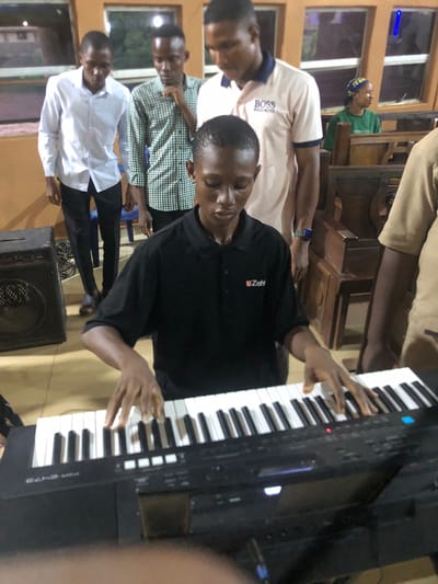 A man plays a keyboard in Nsugbe, Nigeria. Several other men are standing in the background. The interior is dimly lit, with windows showing outdoor views.