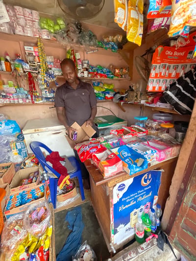 Here is a description of the image:

An overhead shot captures a small shop interior, teeming with products. A man in a brown shirt stands amidst the merchandise, holding a box and examining a small purple package. The shop's walls are lined with shelves stocked with various items, including snack foods like peanuts, Oke Bond glue, and Pure Bliss milk, alongside other goods like water bottles. A small blue plastic chair sits near him, partially covered with a red cloth. Packages of candy labeled "CRASH" are visible. The image likely shows a vendor stocking or organizing his wares in Ado, Nigeria. The presence of "Ramadan Mubarak" signage suggests the photo was taken during or near the Ramadan season. The environment is cramped, with goods stacked high and a general sense of busyness.