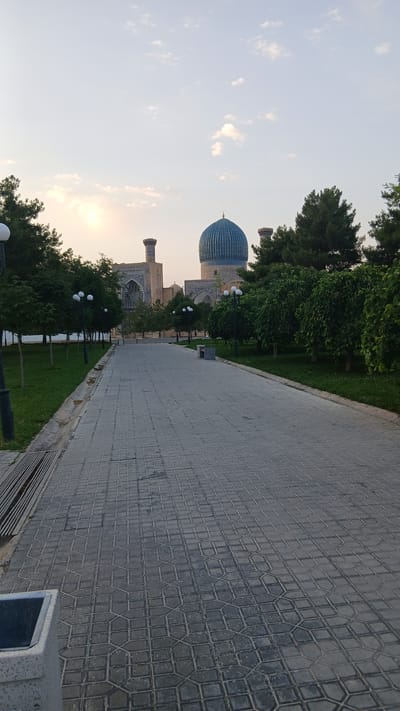 The image shows a paved walkway leading towards a large, ornate building with a prominent blue dome and two minarets. The architecture suggests a historical and possibly religious site. The walkway is made of interlocking gray paving stones arranged in a geometric pattern. On either side of the walkway are manicured green lawns with rows of trees, some of which are flowering or bearing leaves. Several lampposts with multiple globes are spaced along the left side of the walkway. In the background, the building features intricate blue tilework on its facade and dome. The sky is a pale blue with some wispy clouds, and the sun appears to be setting or rising, casting a soft light on the scene. The overall impression is one of serene beauty and historical grandeur. This is likely the Gur-e-Amir Mausoleum in Samarkand, Uzbekistan, given the characteristic architecture.