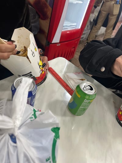 The image shows a table in a restaurant in Segovia, Spain (40.948, -4.118). A person is holding a container of food, likely noodles, with chopsticks. The container is labeled "Asia in Box." There are two soda cans on the table, one blue and one green. The green can is labeled "fuze tea 330 ml." There is a white plastic bag on the table as well. Two people are visible in the image, one holding the container of food and the other seated with their back turned. The environment appears to be a restaurant, with a red refrigerator in the background. There are various objects on the table, including napkins and a red ruler. The person holding the food container is wearing a black shirt and is holding the container with their left hand. The other person is wearing a black jacket and is sitting at the table. The scene is a casual, everyday encounter in a restaurant setting.
