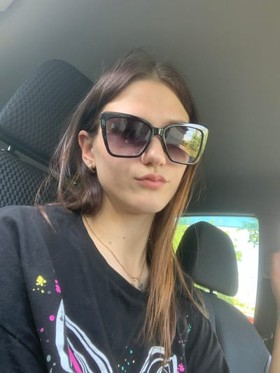 The image shows a young woman in a car, viewed from a slightly elevated, tilted angle. She is wearing large, dark-rimmed sunglasses with a silver accent on the side, which reflect the trees and sky outside. Her long, light brown hair is spread across her shoulders and the seat. She is wearing a black t-shirt with a colorful, abstract graphic design featuring white, pink, and yellow splashes. A delicate gold chain necklace with a small pendant is visible around her neck. The background shows the interior of a car, including a textured black seat and a headrest. Through the car window, greenery and bright daylight are visible, suggesting it is daytime and likely pleasant weather. The woman's pose, with her head tilted and eyes hidden behind sunglasses, could imply a relaxed or casual mood. There is no visible text in the image. The location context is Belogorsk, Russia.