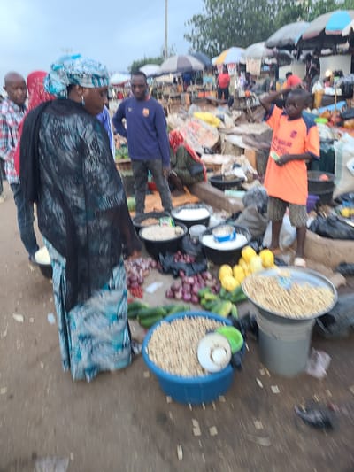This is a vertical, medium-shot, outdoor photograph taken at a busy market. The setting appears to be Nyanya, Nigeria, given the context. A group of people are present, some of whom are involved in commerce. The primary subject in the foreground is a woman wearing a vibrant blue and white headscarf and a dark, patterned garment. Her face is turned away from the camera, suggesting she might be engaged in conversation or tending to a stall. To her left, a man in a red and blue plaid shirt is partially visible, and further left, another person in a red shirt is also present.
In the mid-ground, a young man in an orange t-shirt is looking down, possibly counting money or examining goods. Behind him, a man in a blue long-sleeved shirt stands, also appearing to be interacting with the market environment. The market itself is crowded with various goods and people. Visible objects include large containers of what appear to be grains or nuts, baskets, and produce such as onions and possibly gourds or plantains. There are also sacks and various other items indicative of a bustling marketplace. The ground is dusty, typical of an outdoor market.
The lighting suggests it is daytime, though the exact time is not clear. The sky is overcast, hinting at potentially cloudy or humid weather. The overall atmosphere is one of active trade and daily life in a Nigerian market. No visible text is discernible in the image.
