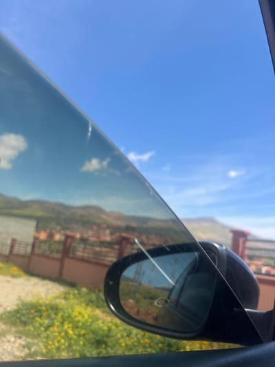 The image shows a view from inside a car, likely traveling through or near the city of Ouaouizert, Morocco. The sky is predominantly blue with a few scattered clouds, visible through the car window. A side mirror is in focus, reflecting the scenery outside, including a blurred view of buildings, possibly houses or shops, and vegetation, likely consisting of plants or grass. The general landscape seems hilly or mountainous, as indicated by the reflected terrain.