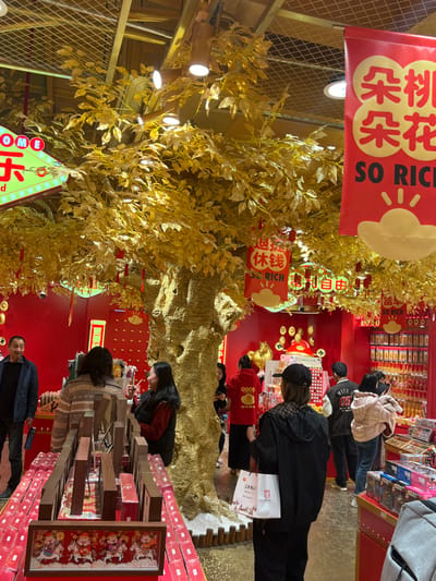 The scene depicts an indoor retail environment located in Shapingba District, China, characterized by a vibrant festive theme. Dominant colors are red for walls and banners, and gold for decorations and a central artificial tree. A large, artificial golden tree with a textured trunk and numerous gold leaves stands prominently in the middle, its branches adorned with red tassel ornaments and small red banner-like decorations bearing Chinese characters such as '退钱' (Tuì qián - refund/money back) and '自由' (Zìyóu - freedom), along with the English phrase 'SO RICH'. The tree's base is surrounded by white material and a low wooden fence. The red walls in the background are decorated with golden cloud motifs and other auspicious symbols. Overhead, structural beams and recessed lighting fixtures are visible. A large red banner hanging from the ceiling displays Chinese characters '朵桃朵花' above 'SO RICH' and a golden sun/cloud graphic. A partially visible neon sign in the upper left reads 'WELCOME'.

Approximately ten individuals are present, casually dressed. In the immediate foreground, a person viewed from behind wears a black jacket and baseball cap, holding a white shopping bag. To their left, a woman makes a peace sign gesture while looking to the right. Other patrons are seen browsing merchandise. On the left, a multi-tiered display unit holds numerous rectangular, reddish-brown packaged items, with a framed image of four cartoon-like figures at its base. Along the right wall, shelves are stocked with various packaged goods, many featuring red and gold packaging. The far back wall displays a large golden stylized pig or ingot decoration and multiple golden coin-like ornaments.