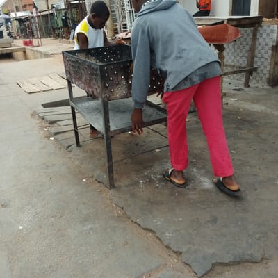 In Tumfure, Nigeria, two young individuals are present. One, wearing a white tank top with yellow trim, is positioned behind a metal grill apparatus, appearing to be engaged with it. The other, clad in a grey hooded jacket and red pants, is facing away from the camera and towards the grill, with their hands near the object. The grill itself is a rectangular, metal structure with a perforated body and appears to be elevated on a stand. The scene is set on a paved outdoor surface, possibly a street or marketplace, with elements of urban infrastructure visible in the background, including buildings with corrugated metal roofs and what appears to be market stalls. The ground is rough and shows signs of wear and debris.